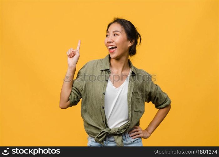 Portrait of young Asian lady smiling with cheerful expression, shows something amazing at blank space in casual clothing and standing isolated over yellow background. Facial expression concept.