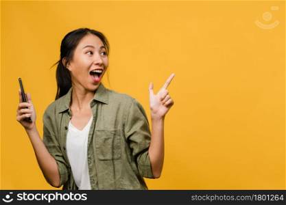 Portrait of young Asian lady using mobile phone with cheerful expression, show something amazing at blank space in casual clothing and stand isolated over yellow background. Facial expression concept.