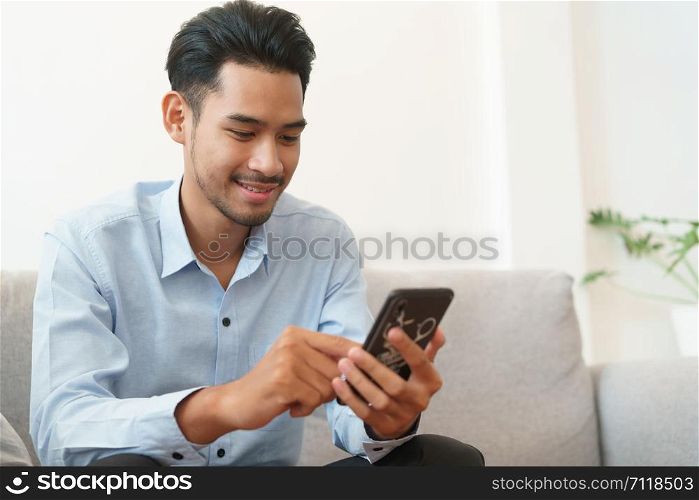 Portrait of young asian man handsome wearing blue shirt is reading using smartphone and sitting on sofa in living room at home
