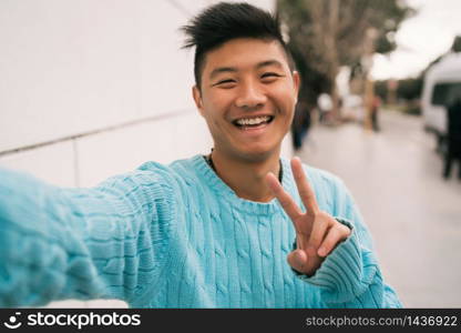 Portrait of young Asian man looking confident and taking a selfie while standing outdoors in the street.