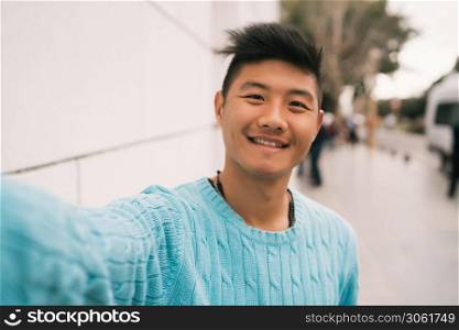 Portrait of young Asian man looking confident and taking a selfie while standing outdoors in the street.