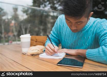 Portrait of young Asian man studying with his digital tablet and taking some notes while sitting in a coffee shop. Technology concept.