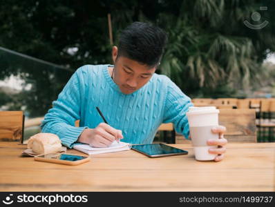Portrait of young Asian man studying with his digital tablet and taking some notes while sitting in a coffee shop. Technology concept.