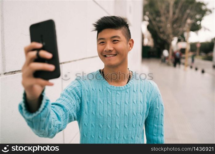 Portrait of young Asian man taking a selfie with his mobile phone while standing outdoors in the street.