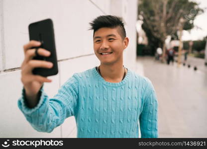 Portrait of young Asian man taking a selfie with his mobile phone while standing outdoors in the street.