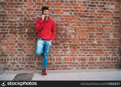 Portrait of young Asian man talking on the phone outdoors against brick wall. Communication concept.