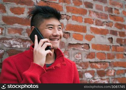 Portrait of young Asian man talking on the phone outdoors against brick wall. Communication concept.