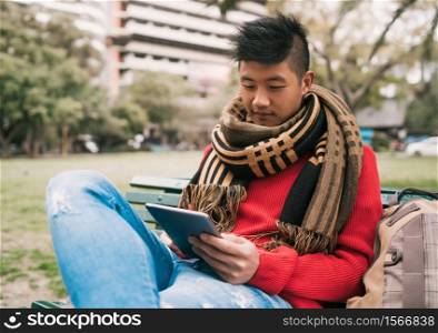 Portrait of young Asian man using his digital tablet while sitting in a bench outdoors. Technology concept.