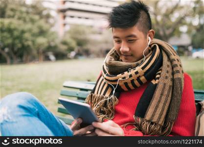 Portrait of young Asian man using his digital tablet with earphones while sitting in a bench outdoors. Technology concept.