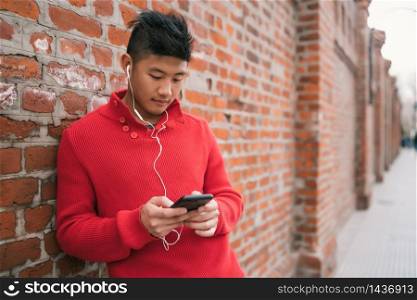 Portrait of young Asian man using his mobile phone with earphones outdoors against brick wall. Communication concept.