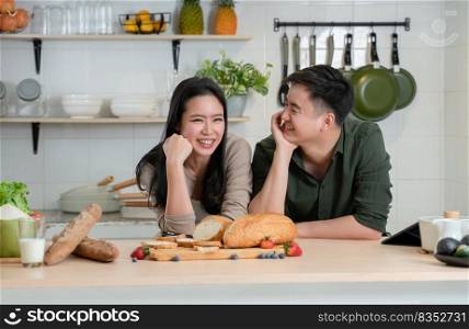 Portrait of young Asian romantic couple smiling, placing hands on chin in the kitchen, happy beautiful wife and handsome husband cooking healthy breakfast food together at home