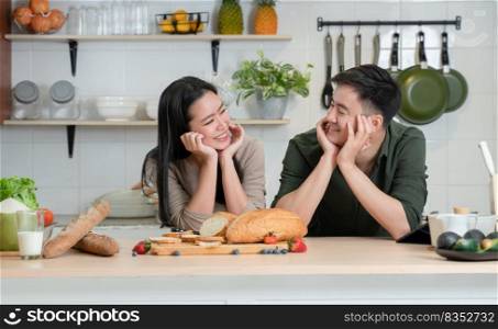 Portrait of young Asian romantic couple smiling, placing hands on chin, looking at each other in the kitchen, happy beautiful wife and handsome husband cooking healthy breakfast food together at home