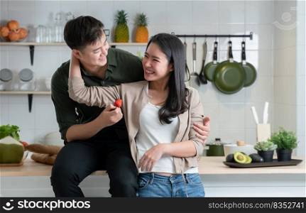 Portrait of young Asian romantic couple standing in the kitchen, happy lover hugging, smiling and looking at each other, handsome man feeding strawberry to beautiful woman while cooking together