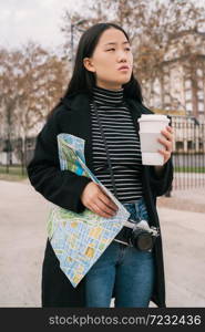 Portrait of young Asian woman holding a map and a cup of coffee outdoors in the street. Travel concept.