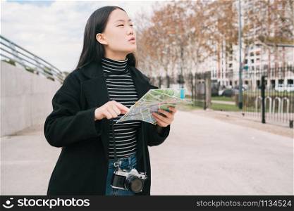 Portrait of young Asian woman holding a map and looking for directions outdoors in the street. Travel concept.