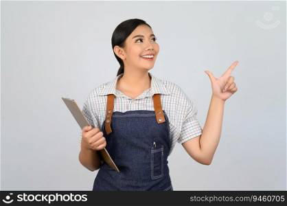 Portrait of young asian woman in waitress uniform holding clipboard and point finger, copy space to insert products for advertisement isolated on white background