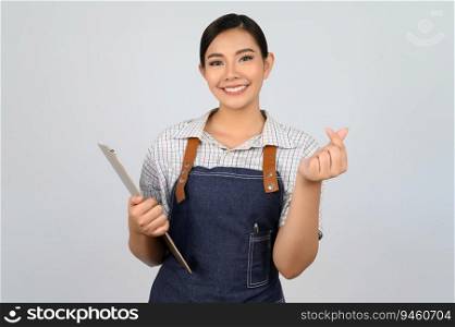 Portrait of young asian woman in waitress uniform holding clipboard and showing mini heart sign pose, copy space to insert products for advertisement isolated on white background