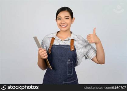 Portrait of young asian woman in waitress uniform holding clipboard and thumb up pose, copy space to insert products for advertisement isolated on white background