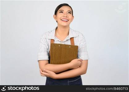 Portrait of young asian woman in waitress uniform holding clipboard on chest with smile and looking up, copy space to insert products for advertisement isolated on white background