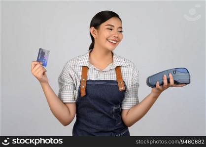 Portrait of young asian woman in waitress uniform pose with credit card and credit card reader machine in hand, copy space to insert products for advertisement isolated on white background