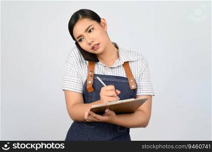 Portrait of young asian woman in waitress uniform talking on smartphone and write order on clipboard during work, copy space for insert products for advertisement isolated on white background