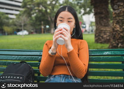 Portrait of young Asian woman listening to music with earphones and drinking a cup of coffee in the park outdoors.