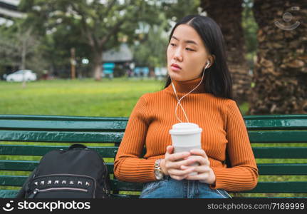 Portrait of young Asian woman listening to music with earphones and holding a cup of coffee in the park outdoors.