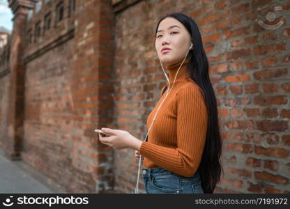 Portrait of young Asian woman listening to music with earphones in the street. Outdoors.