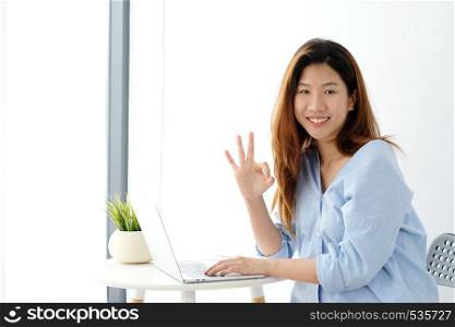 Portrait of young asian woman showing ok hand sign and smiling while working with laptop computer, people with positive gesture