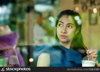 Portrait of young asian woman sitting in coffee shop and holding cup of coffee while looks out the window