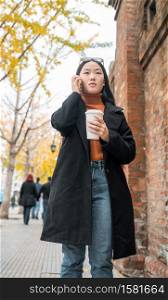 Portrait of young Asian woman talking on the phone while holding a cup of coffee in the street. Urban and communication concept.