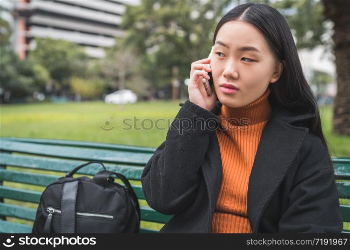 Portrait of young Asian woman talking on the phone while sitting on a bench in the park. Communication concept.