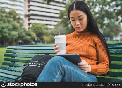 Portrait of young Asian woman using her digital tablet while holding a cup of coffee in the park outdoors.
