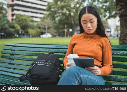 Portrait of young Asian woman using her digital tablet while holding a cup of coffee in the park outdoors.