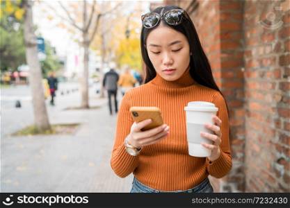 Portrait of young Asian woman using her mobile phone while holding a cup of coffee in the street. Urban and communication concept.