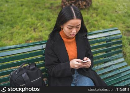 Portrait of young Asian woman using her mobile phone while sitting on a bench in the park. Communication concept.