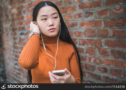 Portrait of young Asian woman using her mobile phone with earphones outdoors in the street. Urban and communication concept.