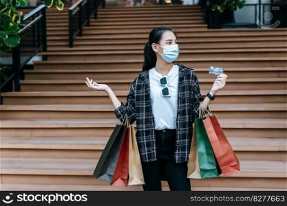 Portrait of young Asian woman wearing protection mask sitting on stairs with shopping paper bag and credit card at shopping mall, New normal Lifestyle and shopping concept
