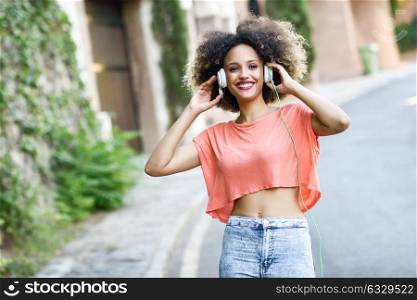 Portrait of young attractive black girl in urban background listening to the music with headphones. Woman wearing orange t-shirt and blue jeans with afro hairstyle