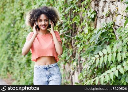 Portrait of young attractive black girl in urban background listening to the music with headphones. Woman wearing orange t-shirt and blue jeans with afro hairstyle
