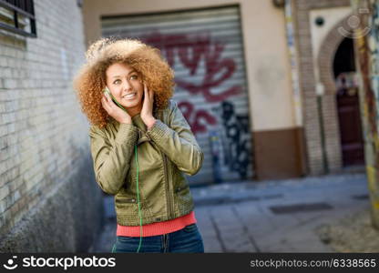 Portrait of young attractive black girl in urban background listening to the music with headphones. Woman wearing leather jacket and blue jeans with afro hairstyle