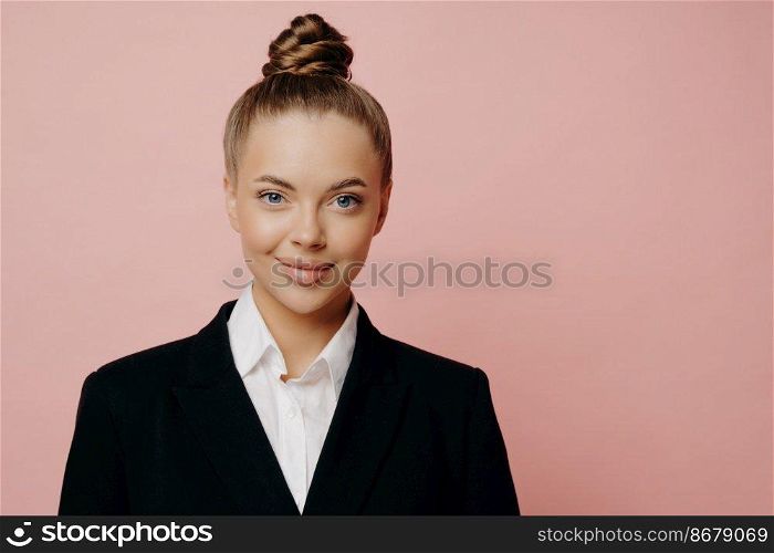 Portrait of young attractive business lady in dark suit with white shirt, hair in bun being happy after talking with sponsors, looking at camera while standing isolated in front of pink background. Happy attractive business lady in dark suit posing in studio