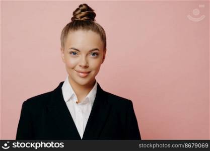 Portrait of young attractive business lady in dark suit with white shirt, hair in bun being happy after talking with sponsors, looking at camera while standing isolated in front of pink background. Happy attractive business lady in dark suit posing in studio