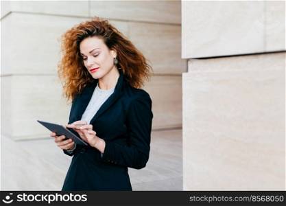 Portrait of young attractive female entrepreneur working on new business project using electronic gadget, having pleased look into screen. Curly woman in black costume standing outdoor near builduing