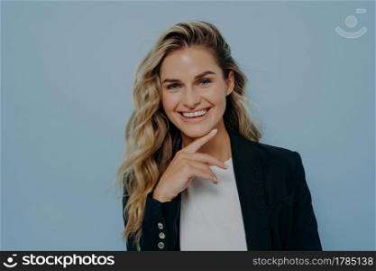 Portrait of young attractive female with finger on her chin showing confidence with gesture and smiling with big toothy smile while posing in studio alone next to blue background. Happiness concept. Attractive blonde woman with finger on her chin smiling at camera