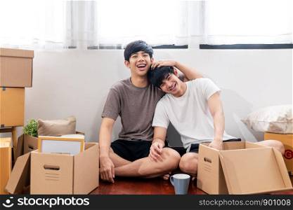Portrait of young attractive happy asian couple man sitting at new home smiling to camera with carton package box storage to move in empty house. LGBTQ relationship and relocating concept