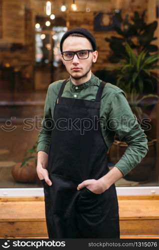 Portrait of young barman in black apron. Coffee house on the background.