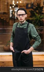 Portrait of young barman in black apron. Coffee house on the background.