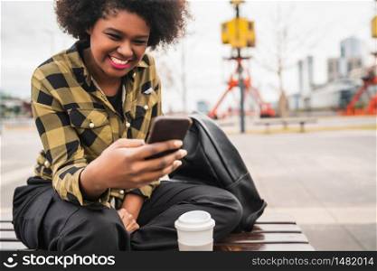 Portrait of young beautiful afro american latin woman using her mobile phone while sitting in a bench outdoors. Communication concept.