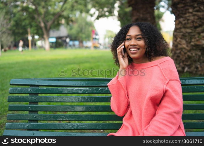 Portrait of young beautiful afro american woman talking on the phone and sitting on a bench in a park. Outdoors.